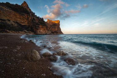 Mavi denizin üzerinde yükselen kaya, batan güneşin turuncu ışınlarıyla mavi gökyüzünün arka planına doğru yükselir.