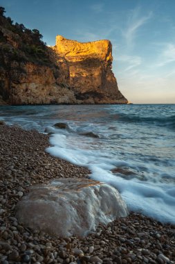 Mavi denizin üzerinde yükselen kaya, batan güneşin turuncu ışınlarıyla mavi gökyüzünün arka planına doğru yükselir.