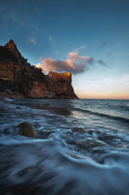 Mavi denizin üzerinde yükselen kaya, batan güneşin turuncu ışınlarıyla mavi gökyüzünün arka planına doğru yükselir.