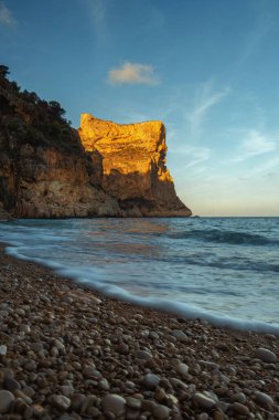 Mavi denizin üzerinde yükselen kaya, batan güneşin turuncu ışınlarıyla mavi gökyüzünün arka planına doğru yükselir.