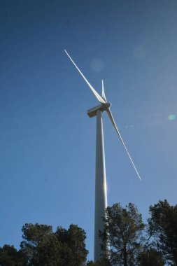Mavi gökyüzüne karşı bir ormanın yeşiliyle çevrili bir rüzgar türbini.