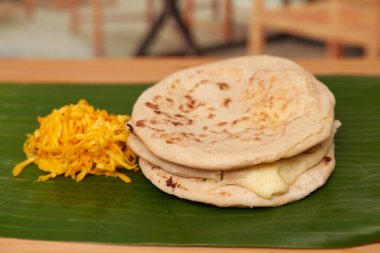 A captivating photo of authentic Salvadoran pupusas, featuring a cheesy filling and culinary artistry, set in a restaurant-style ambiance on a wooden surface accentuated by a leaf clipart