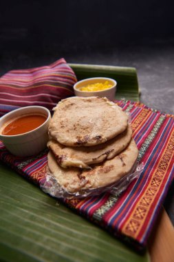 Geleneksel Orta Amerika lezzetlerini vurgulayarak ahşap bir yüzey üzerinde tamale yaprağı üzerinde servis edilen el yazısı ve soslu köpek yavruları..