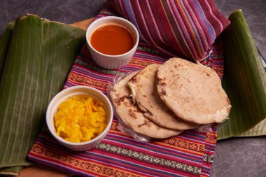 Pupusas with curtido and sauce served on a tamale leaf atop a wooden surface, highlighting traditional Central American flavors. clipart