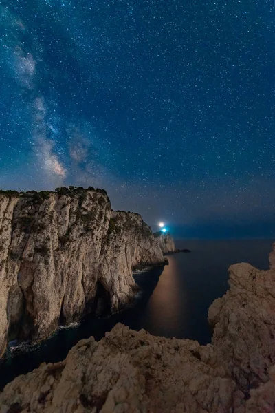 Milky Way and lighthouse on the mountain peak at starry night in summer. Beautiful landscape with cliffs, rocky sea coast, sky with bright milky way and stars. Space and nature. Cape Lefkada, Greece