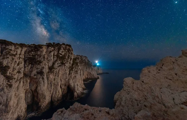 Milky Way and lighthouse on the mountain peak at starry night in summer. Beautiful landscape with cliffs, rocky sea coast, sky with bright milky way and stars. Space and nature. Cape Lefkada, Greece