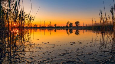 Gölde gün batımı. Sakin su kıyıdaki sazlıkları ve elektrik hatlarını yansıtıyor.