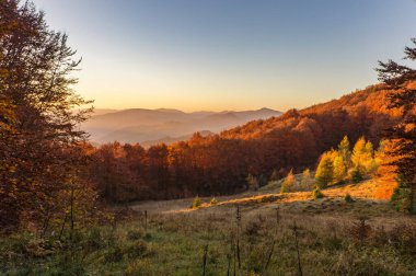 Berrak bir gökyüzü altında canlı yapraklarla kaplı ormanlık tepeleri barındıran nefes kesici bir sonbahar manzarası. Yuvarlanan dağlar ve dağınık bulutlar dingin manzaraya derinlik katıyor.