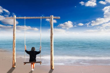 Genç bir kadın güneşli bir günde deniz kenarında salıncakta oturuyor.