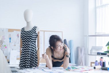 Moda tasarımcısı stüdyoda çalışıyor. Tasarımcı cep telefonu kullanıyor. Asyalı kadın çevrimiçi satıcı telefondaki müşterinin siparişlerini onaylıyor