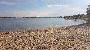 Kumsal, kumlu kıyılar, iskele, binaların yanındaki göl, su, gökyüzü mavisi ve bronz