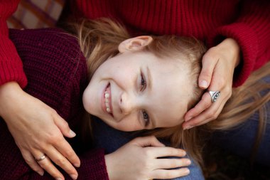 Kafkasyalı neşeli bir kızın, annesinin bacaklarının üzerinde uzanmış sıcak kucaklaşmalarıyla yakın plan portresi. Kadınlar ve kızları sonbahar parkında rahat kırmızı kazaklarla yürüyorlar. Aile görünüşü, mutlu çocukluk.