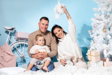 Noel 'de birlikte oturan üç kişinin aile portresi. Mavi arka planda beyaz süslemeler var. Neşeli anne, baba ve küçük oğlu rahat kıyafetler içinde. Yeni yıl, tatil.