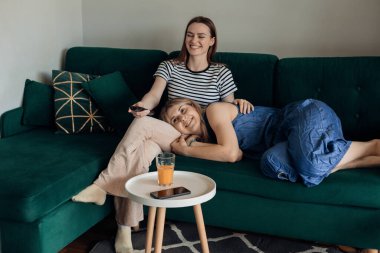 Mutlu genç kadınlar kanepede otururken televizyonda eğlence programı izliyorlar. En iyi arkadaşlar rahat yeşil kanepede rahatlar ve portakal suyu içerler. Gülümse ve TV kumandasında kanal değiştir.