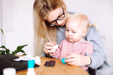 Laptopun yanındaki masada oturan genç bir annenin portresi, haplar, kağıt peçete, kucağında küçük harika bir kız bebek, eve tıbbın nasıl aşılanacağını göstermeye çalışıyor. Hasta zamanı..
