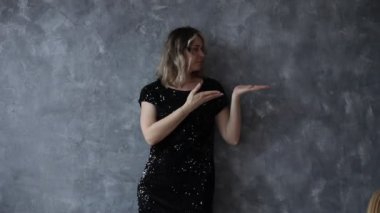 Siyah pullu, neşeli bir kadın. Elleri farklı taraflarda, içi boş gri duvar arkaplanı olan bir elbise. Mesaj için yer. Tatillerde güzel kadınlar giyer. Kutlama, parti, şen şakrak görünüm.