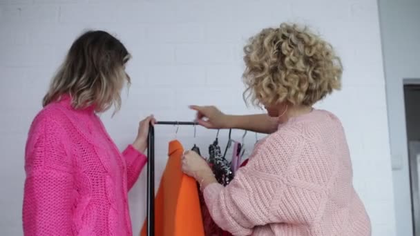 两名身穿粉色针织毛衣的妇女半身站立 在白墙背景下积极选择节日服装 专业的时装设计师提供时尚的橙色夹克 服装选择 — 图库视频影像