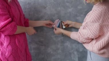 Pembe örülü süveter giyen iki kadın yan yana durur ve gri duvar arkaplanındaki dekorasyon için renk paletini tartışırlar. Profesyonel moda tasarımcısı örnekler sunuyor. Renk seçimi.