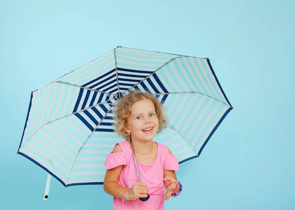 Stock image Cheerful little girl stand under open umbrella on empty blue background, free copy space. Happy child of kindergarten age take shelter from rain. Life insurance and safety conception.