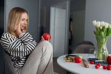 Rahat ev kıyafetleri içinde hamile genç bir sarışın mutfaktaki masada oturuyor ve ellerinde sulu kırmızı bir elma tutuyor. Dengeli beslenme. Hamilelik ve vitaminler. Sağlıklı yaşam tarzı.