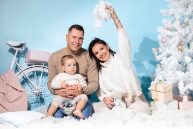 Üç kişilik aile, Noel fotoğraf bölgesinde mavi arka planda beyaz süslemelerle birlikte oturuyorlar. Mutlu anne, baba ve küçük oğul eğlenin ve yapay kar atın. Yeni yıl, tatil, hediye.