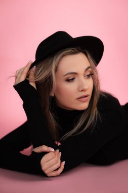 Siyah modaya uygun hippi şapkası ve siyah kazak giyip pembe stüdyo arka planına bakmayan çekici bir kadının dikey görüntüsü. Yüksek moda, güzellik ve kıyafet mağazası
