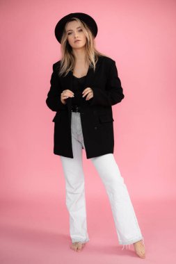 Dikey özgüvenli, modaya uygun, şapkalı ciddi sarışın bir kadın elinde bir şeyle pembe stüdyoda siyah kıyafetler içinde poz veriyor. Model, boş alan, kopyalama alanı ve reklam. Satışlarda en iyi teklif