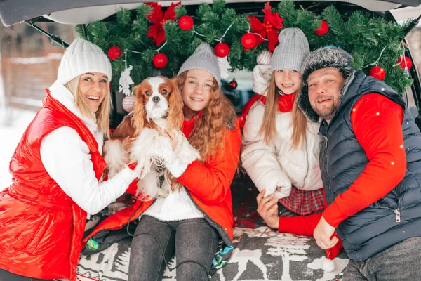 Family party outdoors in Car Trunk among Christmas decoration, mother, father and two daughters are happy to spend time together with dog, red decoration, smiling people in winter