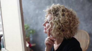 Güzel bir kadın aynanın önünde oturuyor ve arka planda gri duvar manzarası ile akıllanıyor. 50 yaşında bir bayan, şehvetli saç şekillendirme ve makyaj kaldırma zevkini yaşıyor. Yüzün çalışma açısı.