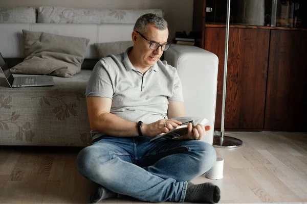 stock image Portrait of concentrated middle-aged man with short hair wearing T-shirt, glasses, jeans, sitting with crossed legs near sofa on floor, holding reading book at home. Spending free time, education.