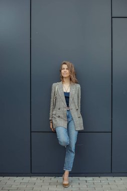 Dikey trend, kendine güvenen, çekici, gri ceketli dalgın sarışın iş kadını mavi duvara yaslanmış, sokakta poz veriyor. Moda, modern kıyafetler ve aksesuarlar. Boşluğu kopyala