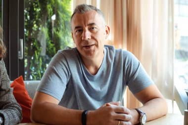 Gri tişörtlü, gri tişörtlü, sırıtan, kendine güveni tam bir adam, kameraya bak, pencerenin yanındaki masaya otur. Mülakat işi, hafif bir ofiste adam toplama. İş toplantısı, insan kaynakları, özgeçmiş ve özgeçmiş