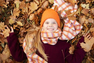Mutlu, neşeli, dişlek, sıcacık elbiseli, güz yaprakları üzerinde uzanan ve renkli yapraklar tutan hoş, küçük sarışın kız. Şükran Günü için dışarıda eğlence ve aktivite. Üst görünüm, tam üstte