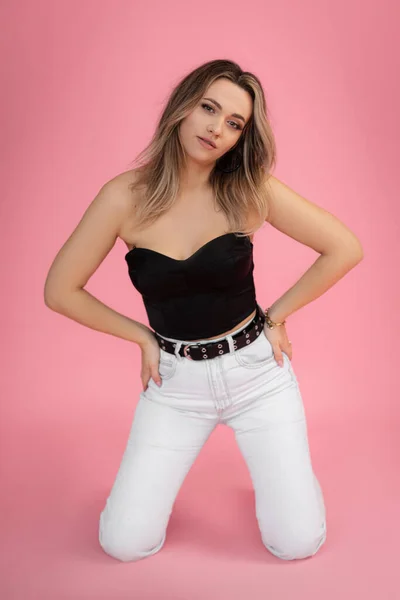 ピンクのスタジオの背景の床に膝の上に立つ流行の黒の上と白のジーンズを身に着けている腰に手を持つ垂直若いブロンドのファッショナブルな 自信のある女性 熱い販売 モックアップ コピースペース — ストック写真