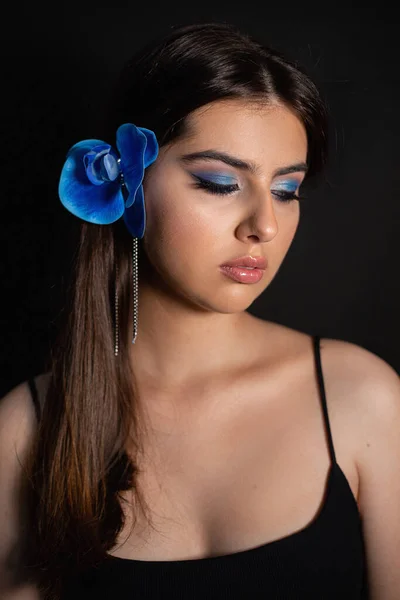 Vertical attractive, serious, gorgeous, romantic brunette woman with make up on perfect skin and blue orchid flowers decoration in hair look down on black background. Perfume industry product