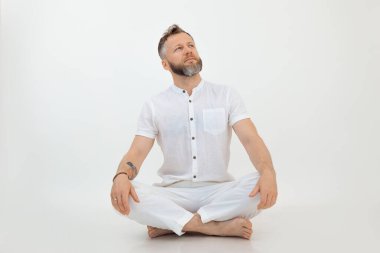 Kendine güvenen, orta yaşlı, kısa gri saçlı, beyaz gömlekli, pantolonlu, bacak bacak bacak üstüne atmış beyaz arka plana bakan orta yaşlı bir adamın portresi. Spor, yoga, eğitim..