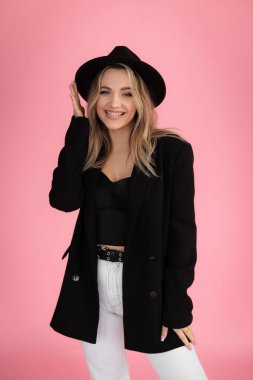 Dikey gülümseme, olumlu, gülen, pembe stüdyoda siyah kıyafetli parlak tenli, kendine güvenen sarışın kadın. Moda mankeni, magazin için poz veren şık genç bir kadın. Boşluğu kopyala