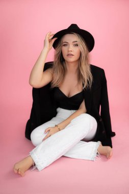 Dikey genç, yalın ayak, baştan çıkarıcı, güzel, çekici, uzun sarı saçlı, siyah şapkalı kadın, pembe arka planda bacak bacak üstüne atarak yerde oturuyor. Mükemmel cilt için kozmetoloji. Boşluğu kopyala