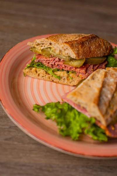 Vertical shot of pink plate with tasty, crispy sandwich with meat and cucumbers, parsley, split in two pieces on wooden table. Healthy subway snack and new recipe of cafe. Delivery order and service
