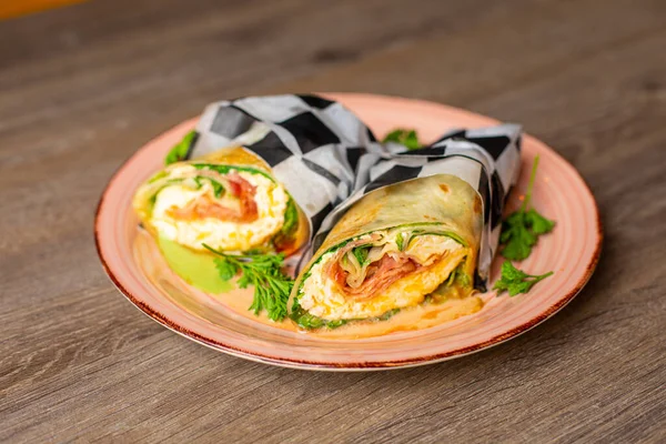Appetizing bacon rolls stuffed with greenery, sauce and ham wrapped in checkered napkin, served on plate on wooden table. New cafe menu, advertising of season snacks, fast food and junk food.