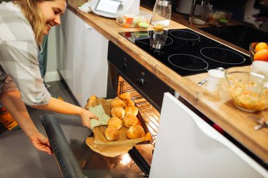 Kırpılmış gülen sarışın kadın mutfaktaki indüksiyon fırınından çörek tepsisi çıkartıyor. Hobi yapmak ve evde hamur işi hazırlamak. Tatillerde ya da hafta sonlarında lezzetli çeşitlilik