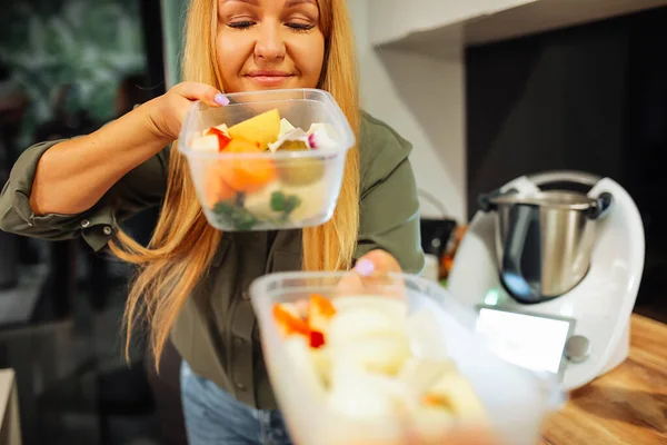 Joyful blond mature woman carry and smelling fresh vegetables and fruit in plastic containers near blender in wooden kitchen corner. No smell, organic food preservation. Fresh food delivery, takeaway