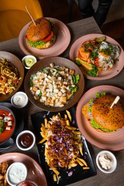 Kafedeki ahşap masada çeşitli lezzetli abur cuburların dikey fotoğrafı. Bbq burger, Cesar salatası ve kızarmış ekmek, makarna, sos, patates kızartması ve et, kızarmış yumurtalı sandviç pizza.