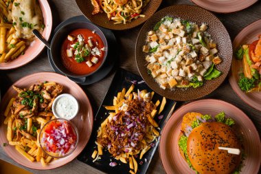 Kafedeki ahşap masada ızgara fast food. Ekmek arası kızarmış domates çorbası, sos, hamburger, patates kızartması, makarna, sandviç ve Sezar salatası. Yeni malzemeler, restoranda sezon menüsü. Üst görünüm