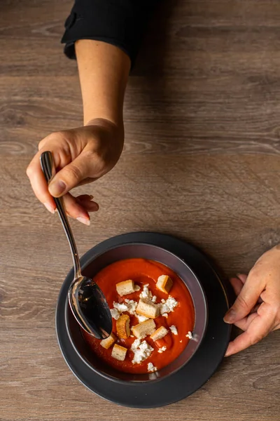 Vertical photo of cropped woman hands stir red tomato cream soup with soup crouton toasts on black fashionable plate on wooden table in cafe. Vegan organic diet and modern service in restaurant