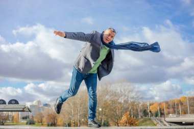 Kısa saçlı, ceketli, uçan mavi atkı takan, gözlüklü, kot pantolonlu, beton korkulukta tek ayak üzerinde duran, sonbaharda güneşli bir günde şehirde elini uzatan orta yaşlı bir adamın portresi..