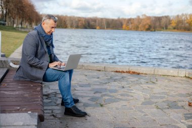 Kısa gri saçlı, ceketli, mavi atkılı, gözlüklü, kot pantolonlu orta yaşlı bir iş adamının portresi. Sonbaharda şehirde, parkta, ahşap bankta oturuyor ve dizüstü bilgisayarla yazı yazıyor..