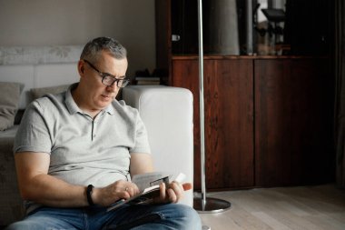 Kısa saçlı, gri tişörtlü, gözlüklü, kot pantolonlu, koltuğun yanında oturmuş kitap okuyan orta yaşlı bir adamın portresi. Boş zaman geçirmek, hobi, eğitim.