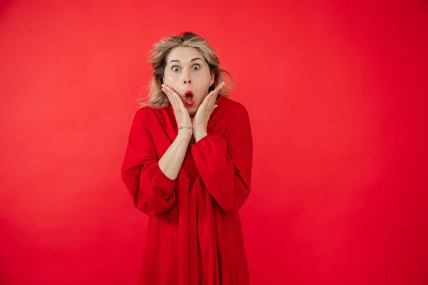 Portrait of gorgeous young woman in bright red dress wondering with open mouth and bulging eyes isolated on red background. Wow effect, great result, surprising outcome. Fashion and beauty industry.