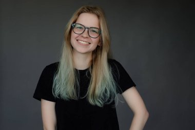 Uzun sarı saçlı, gözlüklü, siyah tişörtlü, gri arka planda kameraya bakan genç ve güzel bir bayan öğrencinin portresi. Stüdyo. Güzellik, moda, gençlik.